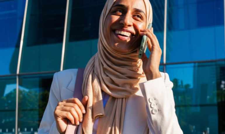 Confident professional woman in beige hijab smiling while on a phone call outside a modern office building – representing career success in mid-2026 | Radeya Global